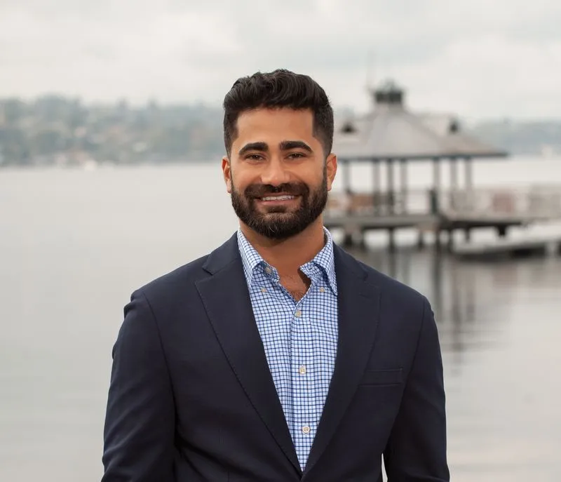 A man in a dark suit and checked shirt stands by a lakeside pier with trees and cloudy skies in the background.