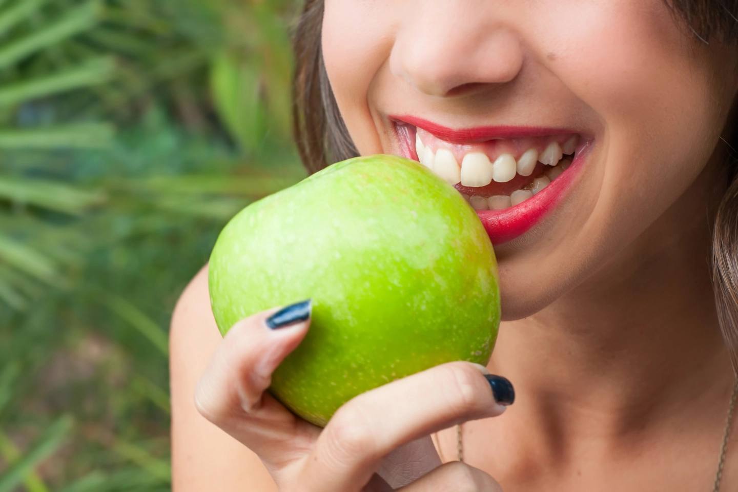 A woman is smiling while holding a green apple close to her mouth, showcasing her bright, healthy teeth.