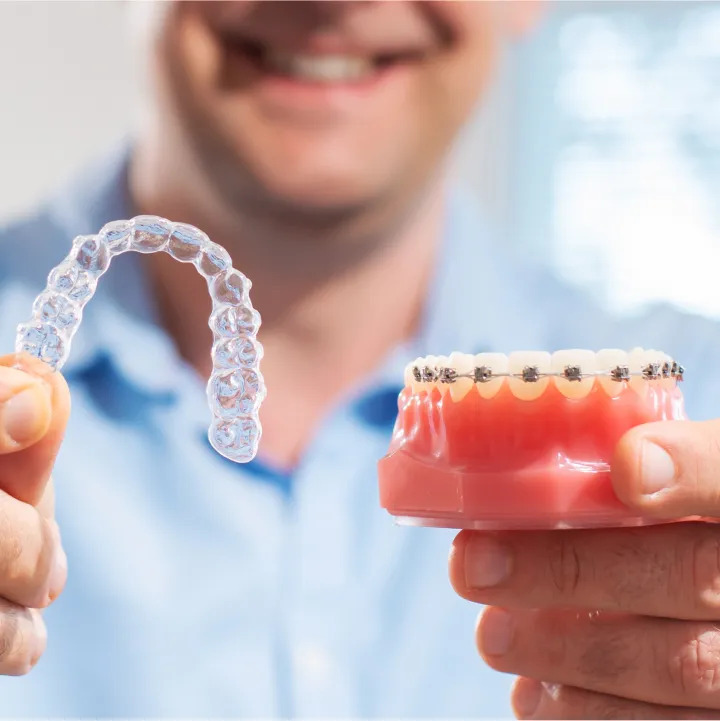 A dentist holding a clear dental aligner in one hand and a model of metal-braced teeth in the other hand.