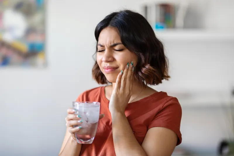 A woman holding a glass of ice water grimaces in pain while touching her jaw, indicating a dental issue.
