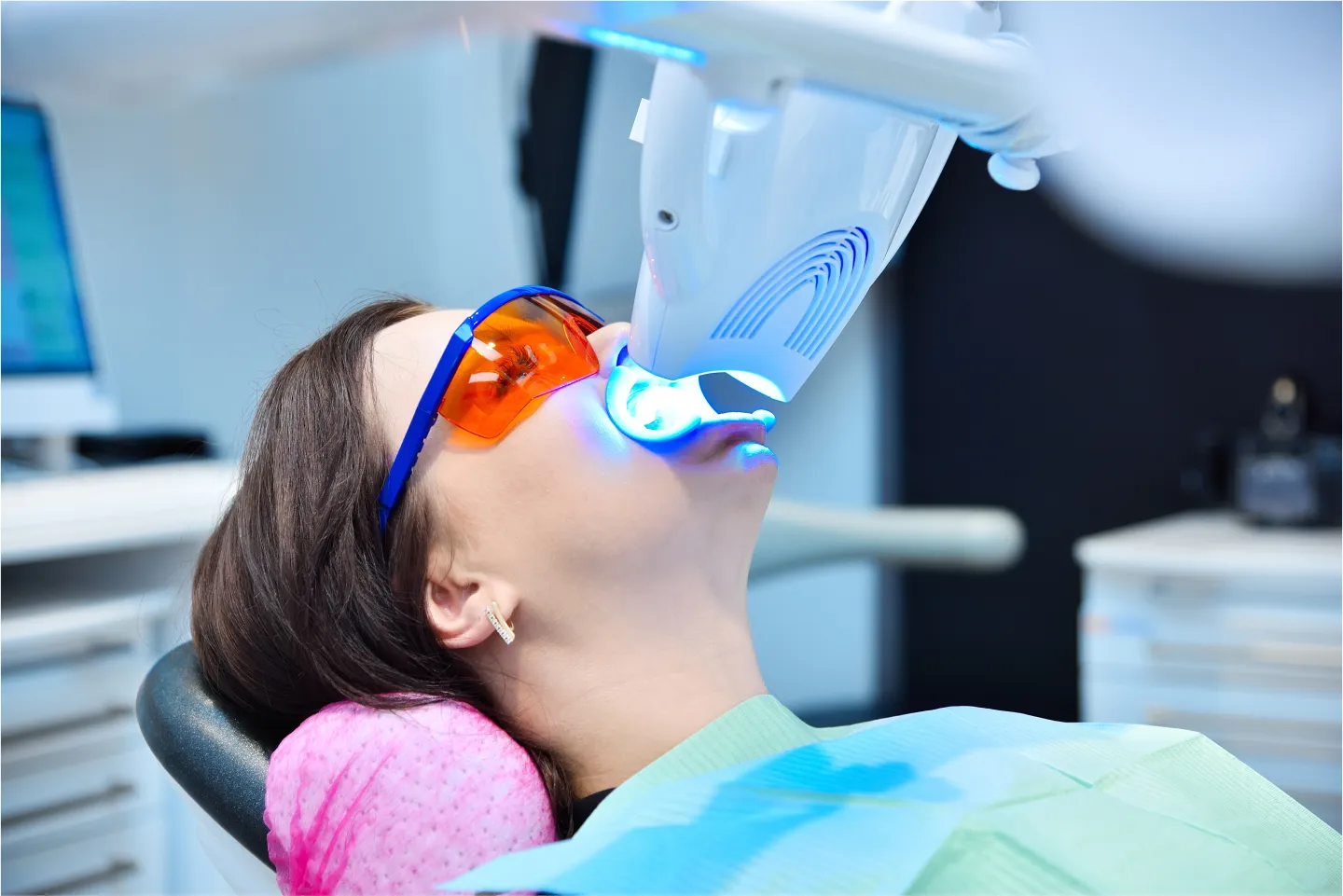 A patient receiving teeth whitening treatment with a blue LED light in a dental office.