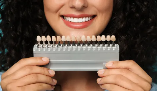 A woman with curly hair smiling while holding a dental shade guide for teeth color matching at SmileVibe Dentistry.