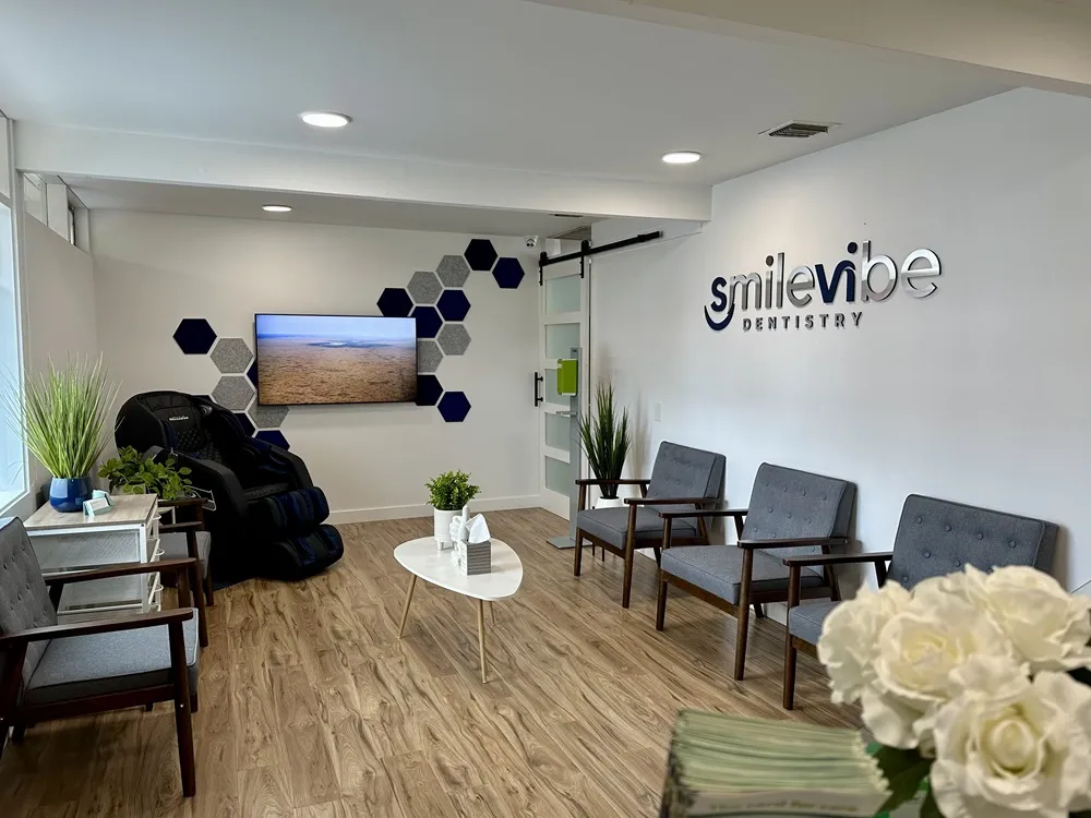 Waiting area at SmileVibe Dentistry featuring gray chairs, a TV displaying a landscape, plants, and a decorative wall.