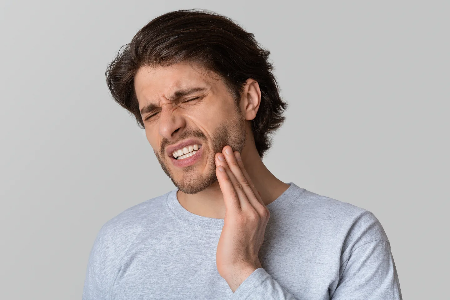 A man with shoulder-length hair is grimacing and holding his jaw, indicating a toothache or dental pain.