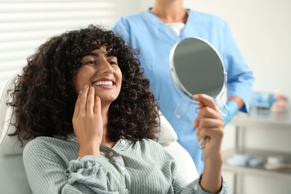 A patient smiling and touching her face while looking at her reflection in a mirror during a dental appointment.