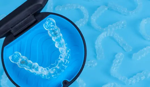Clear dental aligners are displayed in a black case with a blue interior, surrounded by additional aligners on a blue background.