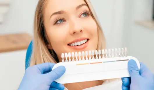 A patient smiles while a dentist holds a dental shade guide to compare tooth colors in a dental office.