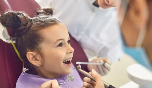 A young girl smiling in a dental chair while a dentist examines her teeth using dental tools.